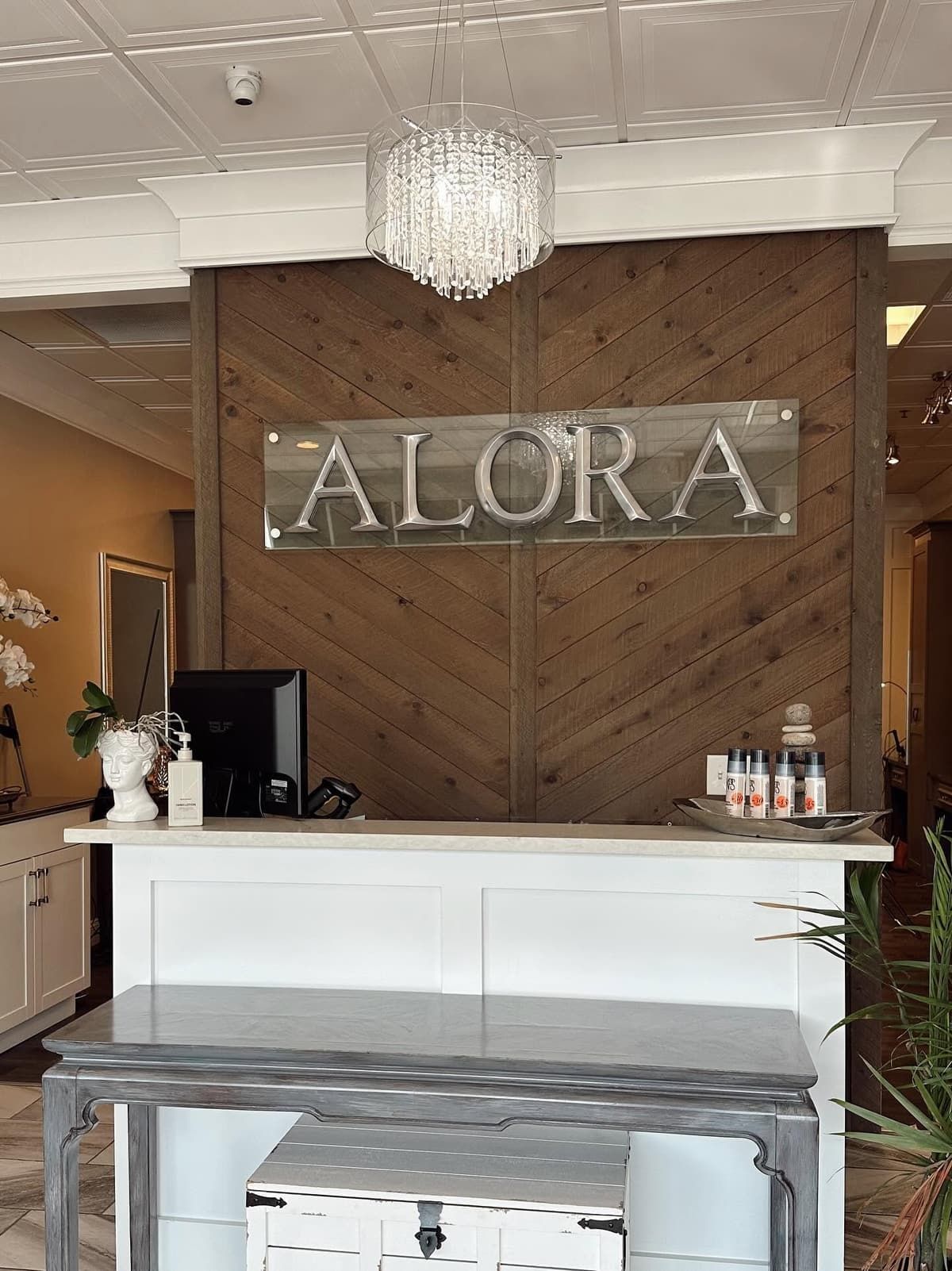 The reception desk at Alora Salon & Wellness Spa in Robinson Township, with the ALORA sign on a chevron wood wall and a crystal chandelier